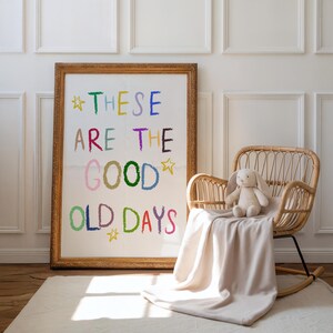 May include: A framed print with the text "THESE ARE THE GOOD OLD DAYS" in colorful letters. A plush bunny sits on a wicker rocking chair with a beige blanket. The room has white paneling and a light-colored rug.