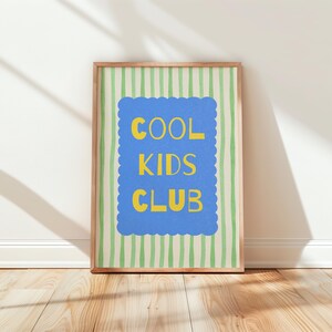 K&ouml;nnte beinhalten: Ein gerahmter Druck mit den Worten "COOL KIDS CLUB" in Gelb auf einem blauen, wellenf&ouml;rmigen Hintergrund. Der Hintergrund hat vertikale gr&uuml;ne und wei&szlig;e Streifen. Der Rahmen ist hellbraun. Der Druck lehnt an einer wei&szlig;en Wand auf einem Holzboden.
