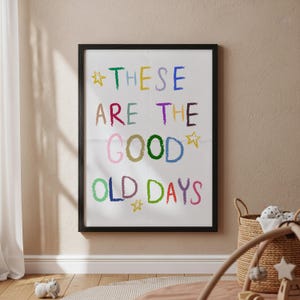 May include: A framed print with the words "THESE ARE THE GOOD OLD DAYS" in colorful, crayon-style lettering. The print is in a black frame and hangs on a beige wall. A woven basket and a baby play gym are in the foreground.