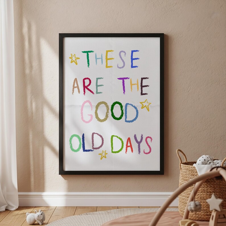 May include: A framed print with the words "THESE ARE THE GOOD OLD DAYS" in colorful, crayon-style lettering. The print is in a black frame and hangs on a beige wall. A woven basket and a baby play gym are in the foreground.