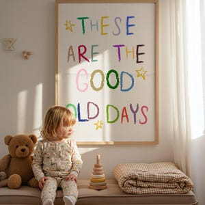 May include: A child in a daisy-print onesie sits on a bench next to a teddy bear and a wooden stacking toy. A framed artwork with the text "THESE ARE THE GOOD OLD DAYS" hangs on the wall.