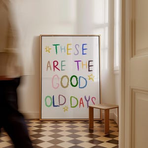 Pode incluir: Uma impressão de arte emoldurada com as palavras "THESE ARE THE GOOD OLD DAYS" em letras coloridas e manuscritas. A impressão está em uma moldura dourada e é exibida em uma sala com um pequeno banco de madeira e um chão xadrez.