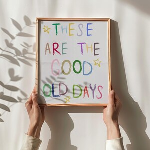 May include: A framed print with the words "THESE ARE THE GOOD OLD DAYS" in colorful, handwritten-style lettering. The print is held by two hands against a white wall, with a shadow of leaves in the background.