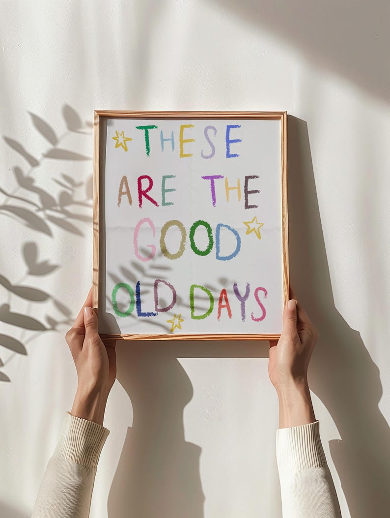 May include: A framed print with the words "THESE ARE THE GOOD OLD DAYS" in colorful, handwritten-style lettering. The print is held by two hands against a white wall, with a shadow of leaves in the background.