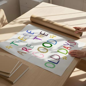 May include: A white poster with the text "THESE ARE THE GOOD OLD DAYS" in colorful crayon-style lettering. A rolled-up brown paper tube and several brown notebooks are on a light wood table.