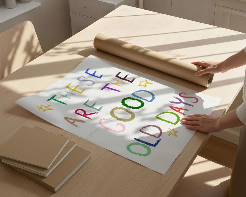 May include: A white poster with the text "THESE ARE THE GOOD OLD DAYS" in colorful crayon-style lettering. A rolled-up brown paper tube and several brown notebooks are on a light wood table.