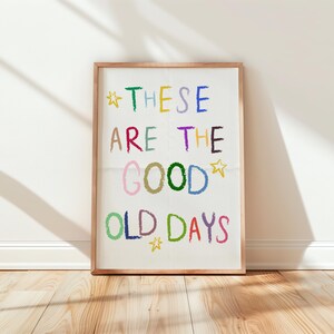 May include: A framed art print with the words "THESE ARE THE GOOD OLD DAYS" in colorful, crayon-style lettering. The print is in a light wood frame and leans against a white wall on a wooden floor. The text includes stars.