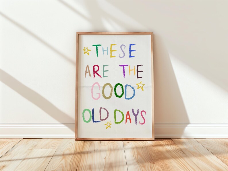 May include: A framed art print with the words "THESE ARE THE GOOD OLD DAYS" in colorful, crayon-style lettering. The print is in a light wood frame and leans against a white wall on a wooden floor. The text includes stars.