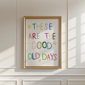 May include: A framed art print with the words "THESE ARE THE GOOD OLD DAYS" in colorful, crayon-style lettering. The text is set against a white background, framed in a gold-colored frame, and accented with star shapes.
