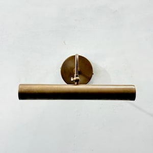 May include: A wall-mounted picture light fixture with a cylindrical horizontal shade and a circular backplate. The entire piece is finished in a brushed antique brass metal, featuring an adjustable arm with a small tightening knob for positioning.