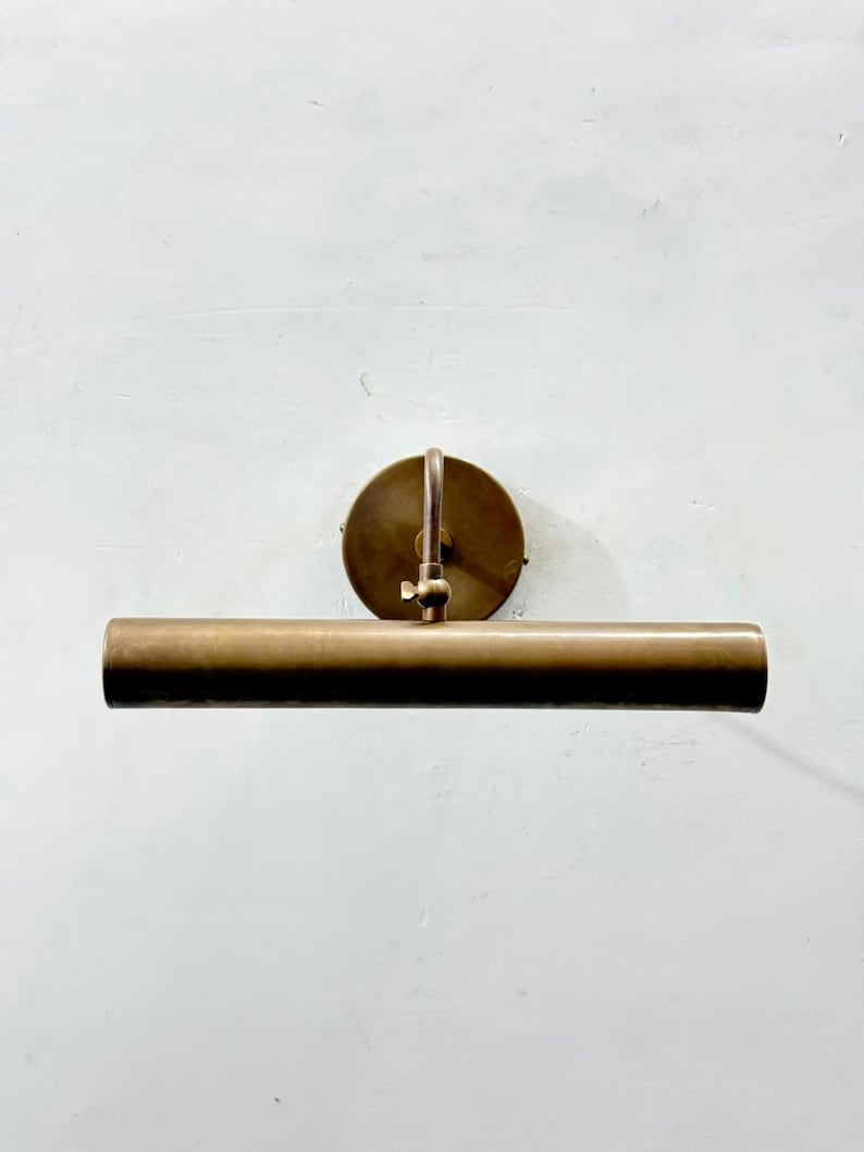 May include: A wall-mounted picture light fixture with a cylindrical horizontal shade and a circular backplate. The entire piece is finished in a brushed antique brass metal, featuring an adjustable arm with a small tightening knob for positioning.
