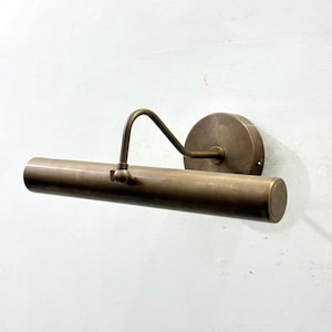 May include: A wall-mounted picture light with a cylindrical brass lamp head and a circular mounting plate. The fixture features an adjustable arm that connects the horizontal tube to the base, finished in a muted, brushed metallic tone.