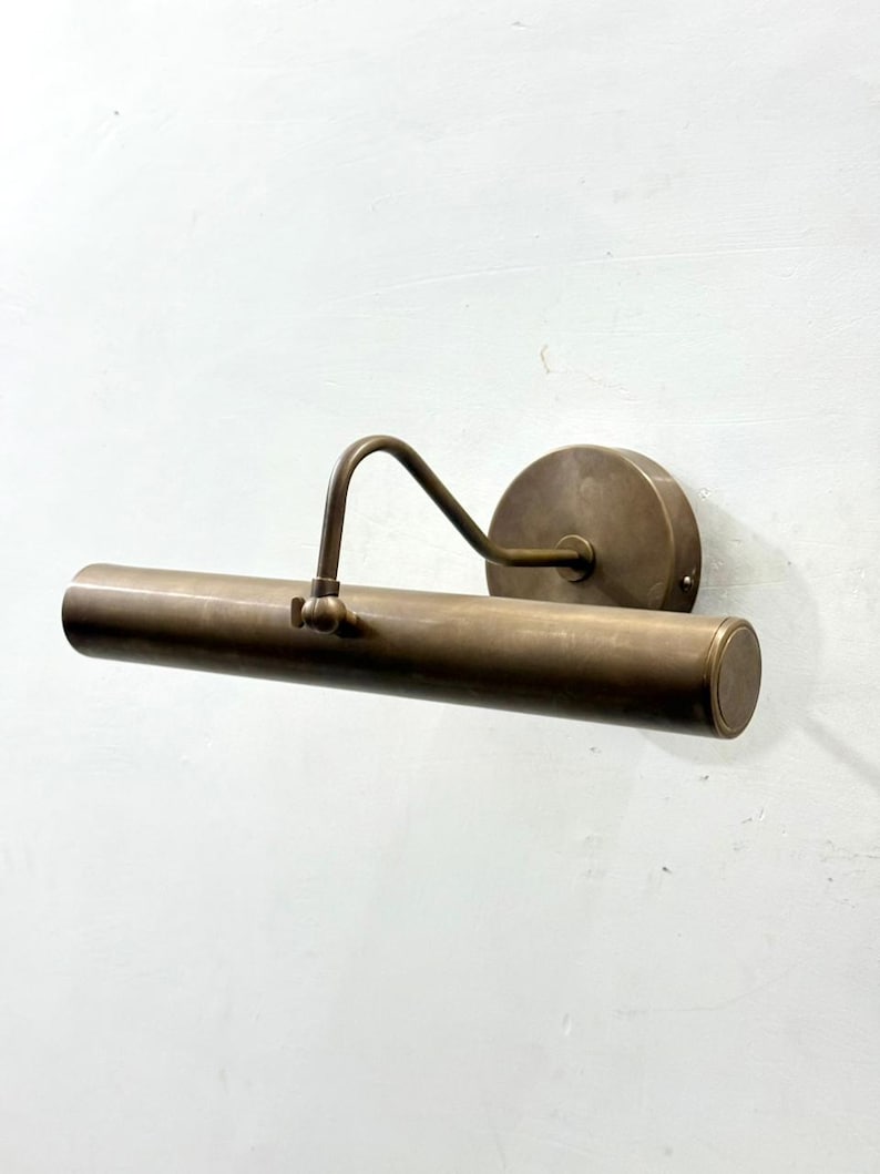 May include: A wall-mounted picture light with a cylindrical brass lamp head and a circular mounting plate. The fixture features an adjustable arm that connects the horizontal tube to the base, finished in a muted, brushed metallic tone.