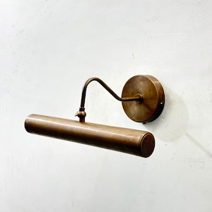 May include: A vintage-style picture light fixture with a bronze finish. The light has a cylindrical shade and a curved arm connecting to a round wall mount. The fixture is mounted on a white wall.