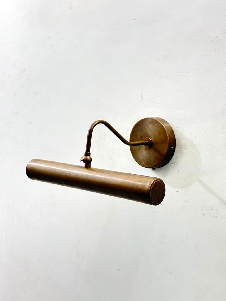 May include: A wall-mounted picture light with an antique brass finish. The fixture features a circular base plate, a curved arm, and a horizontal cylindrical light housing. The design includes a small adjustment knob on the top of the cylinder.