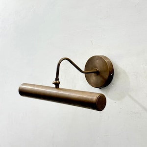 May include: A wall-mounted picture light fixture with a cylindrical head and a curved arm, finished in a matte antique brass tone. The fixture features a circular base plate that attaches to the wall, supporting the horizontal light tube.