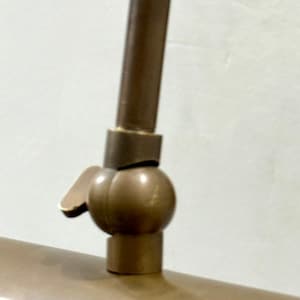 May include: A metallic bronze-toned lamp joint featuring a spherical swivel mechanism with a small adjustment lever. The component connects a vertical rod to a horizontal cylindrical lamp shade, showcasing a smooth, matte finish throughout.