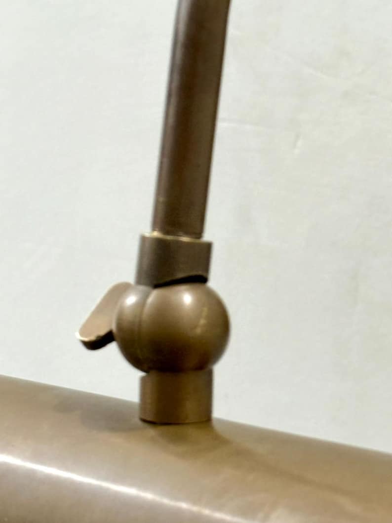 May include: A metallic bronze-toned lamp joint featuring a spherical swivel mechanism with a small adjustment lever. The component connects a vertical rod to a horizontal cylindrical lamp shade, showcasing a smooth, matte finish throughout.