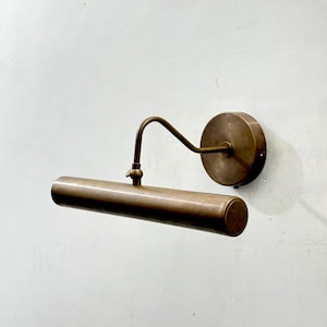 May include: A wall-mounted picture light with an antique brass finish. The fixture features a circular wall plate, a curved arm, and a horizontal cylindrical light housing. The design includes a small adjustable joint connecting the arm to the tube.