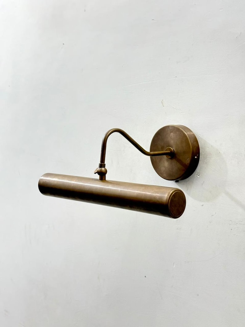 May include: A wall-mounted picture light with an antique brass finish. The fixture features a circular wall plate, a curved arm, and a horizontal cylindrical light housing. The design includes a small adjustable joint connecting the arm to the tube.