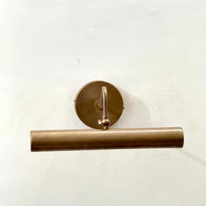 May include: A wall-mounted picture light with a cylindrical brass-toned metal shade and a circular mounting plate. The fixture features an adjustable arm connected to the center of the shade, allowing for directional light positioning on a wall.