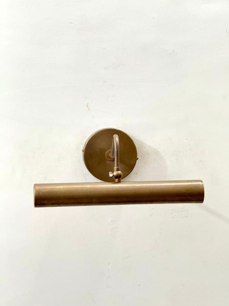 May include: A wall-mounted picture light with a cylindrical brass-toned metal shade and a circular mounting plate. The fixture features an adjustable arm connected to the center of the shade, allowing for directional light positioning on a wall.