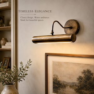 May include: A wall-mounted picture light with an antique brass finish, featuring a cylindrical lamp head attached to a circular base by a curved arm. The light emits a warm glow downward. Text reads: TIMELESS ELEGANCE. Classic design. Warm ambiance. Made for beautiful spaces.