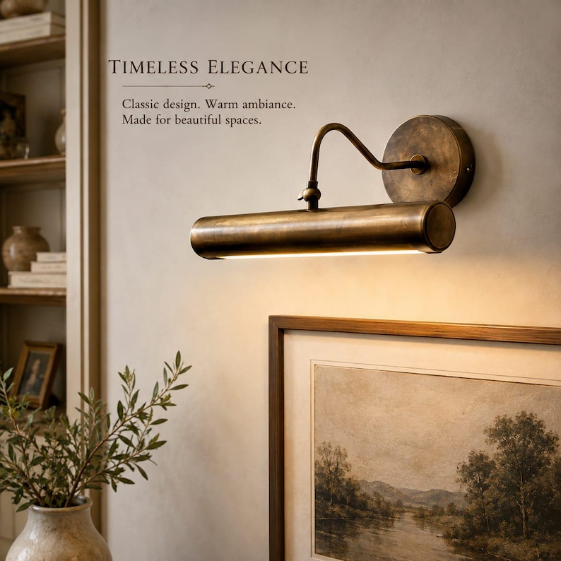 May include: A wall-mounted picture light with an antique brass finish, featuring a cylindrical lamp head attached to a circular base by a curved arm. The light emits a warm glow downward. Text reads: TIMELESS ELEGANCE. Classic design. Warm ambiance. Made for beautiful spaces.