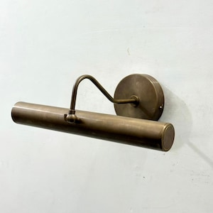May include: A wall-mounted picture light with a cylindrical brass finish lamp head. The fixture features a circular wall plate and a curved arm that connects to the center of the horizontal tube, allowing for adjustable positioning of the light source.