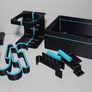 May include: A collection of black and teal 3D-printed organizers and accessories. Includes a box, hooks, a cup holder, and other items. The box features a mountain and tree design. The items have teal accents.