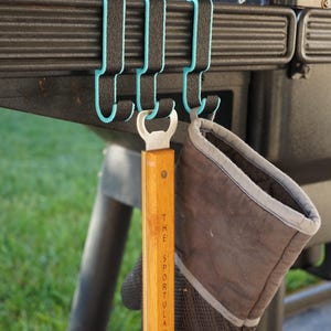 May include: Black grill hooks with a teal accent, holding a wooden bottle opener and a gray oven mitt. The bottle opener has the text "THE SPORTULA" printed on it. The items are hanging on a black grill.