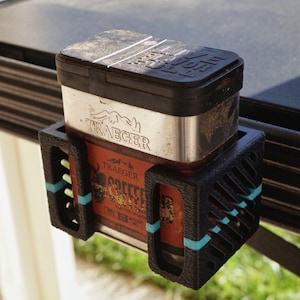 May include: A black 3D-printed spice holder with teal accents, holding a Traeger spice tin. The spice tin is silver with a black lid and a red label. The holder is mounted on a black railing, suitable for outdoor grilling.
