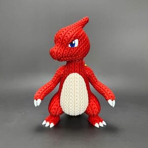 Puede incluir: Una figura roja tejida con vientre color crema, ojos azules y detalles blancos en pies y garras. La figura tiene un cuerno en la cabeza y está en posición vertical. El artículo es una artesanía hecha a mano.
