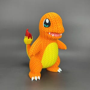 Puede incluir: Figura de Charmander naranja tejida con vientre amarillo y garras blancas. La figura tiene una llama roja en la cola, una boca sonriente y ojos grandes azules y blancos. Personaje popular de la franquicia Pokémon.