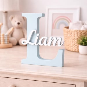 Puede incluir: Una letra "L" de madera azul claro con el nombre "Liam" en escritura blanca. La pieza decorativa se exhibe sobre una superficie de madera clara, ideal para una habitación infantil.