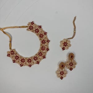 May include: A gold-toned jewelry set featuring a necklace, earrings, and a headpiece. The necklace and earrings have a floral design with red and clear accents. The headpiece has a similar floral design. The set is displayed on a white surface.