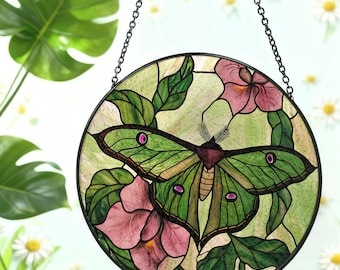 May include: Round stained glass window featuring a green moth with pink accents, surrounded by pink flowers and green leaves. The artwork is suspended by a black chain. The design is set against a light green background.