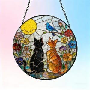 May include: Round stained glass window art featuring two cats gazing at a blue bird perched on a branch. The scene includes a yellow sun, colorful flowers, and a black metal chain for hanging. The cats are black and orange.