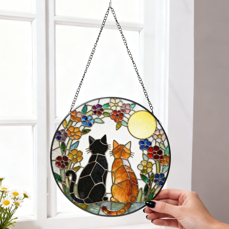 May include: Stained glass round window hanging featuring two cats, one black and one orange, gazing at a yellow moon. The cats are surrounded by colorful flowers. The piece is suspended by a chain.