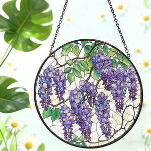 Pode incluir: Uma janela redonda de vitral com um design de glicínias. A obra de arte apresenta flores de glicínias roxas em cascata, folhas verdes e galhos marrons sobre um fundo branco. A janela é emoldurada em preto e suspensa por uma corrente.