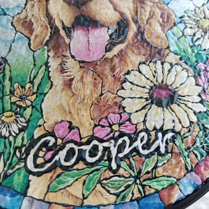 May include: A round, decorative garden stone featuring a Golden Retriever dog with its tongue out, surrounded by colorful flowers. The name "Cooper" is written in white script. The stone has a mosaic-like border.