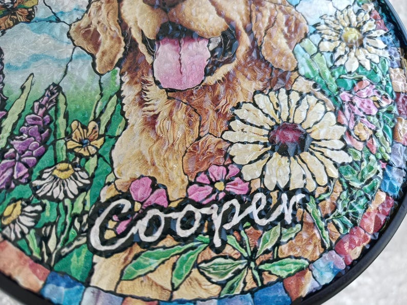 May include: A round, decorative garden stone featuring a Golden Retriever dog with its tongue out, surrounded by colorful flowers. The name "Cooper" is written in white script. The stone has a mosaic-like border.