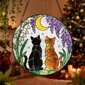 May include: A round stained glass window featuring two cats gazing at a crescent moon. The cats are black and orange, set against a backdrop of purple wisteria, white flowers, and green foliage. The window is hanging from a chain.