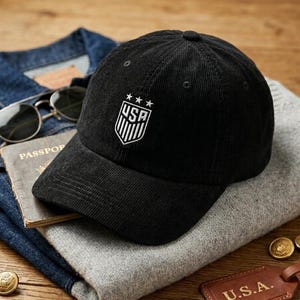 May include: Black corduroy baseball cap with the USA emblem embroidered on the front. The cap has a curved brim and a structured crown. The hat is displayed on a gray sweater, with sunglasses and a passport in the background.