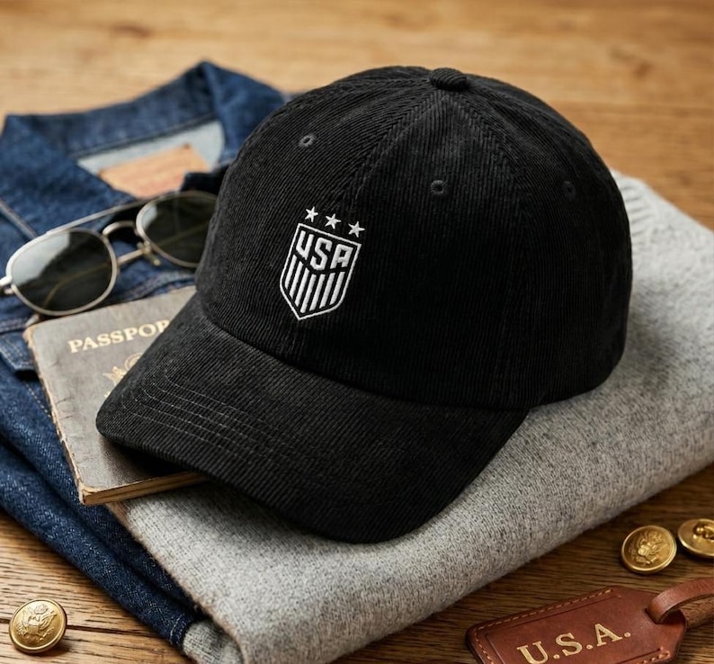 May include: Black corduroy baseball cap with the USA emblem embroidered on the front. The cap has a curved brim and a structured crown. The hat is displayed on a gray sweater, with sunglasses and a passport in the background.
