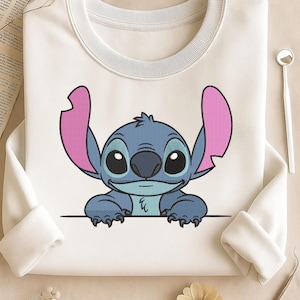 May include: White sweatshirt featuring a blue cartoon character with large ears and a friendly expression. The character has large black eyes, a dark blue nose, and pink inner ears. The design is centered on the sweatshirt.
