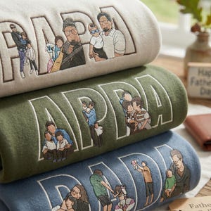 Op de afbeelding: Een stapel van drie geborduurde sweatshirts in crème, groen en blauw. Elk sweatshirt heeft het woord "PAPA" of "DAD" in grote witte letters, met geborduurde familieportretten. De sweatshirts zijn een Vaderdagcadeau.