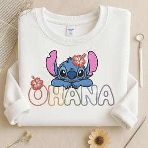 May include: White crew neck sweatshirt with a blue cartoon character with large ears and a red flower. The word "OHANA" is embroidered in colourful letters below the character. A fun and playful design.