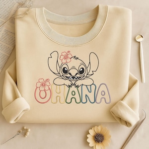 Peut inclure: Sweat-shirt crème avec une illustration de dessin animé de Stitch avec une fleur rose et le mot "OHANA" en couleurs arc-en-ciel. Le sweat-shirt a un col rond et des manches longues.