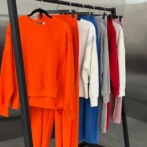 Pode incluir: Um cabideiro exibe vários conjuntos de camisolas e calças de fato de treino a condizer em várias cores. Os conjuntos incluem laranja, azul, branco, cinzento, vermelho e rosa. Cada conjunto apresenta uma camisola de gola redonda e calças de fato de treino a condizer.